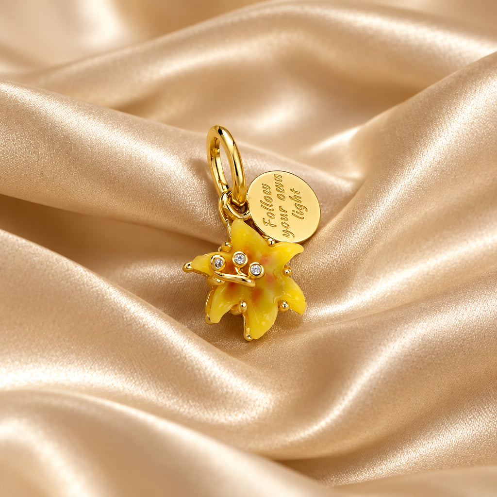 A gold charm pendant featuring a yellow and orange enamel flower with small rhinestones, accompanied by a round engraved gold disc charm, displayed on a beige satin fabric background.
